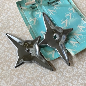 May include: Two chrome-plated star-shaped decorative items with pointed edges and a central hole. The items are displayed on a patterned beige surface, with a partially visible package in the background. The package has the text "Amerock" and "Genuine".