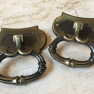 Op de afbeelding: Twee antieke messing ladegrepen met een decoratieve ovale ring en een haakvormige handgreep. De grepen hebben een donkerbruine houten inzet.