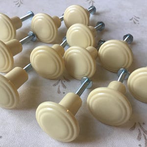 Peut inclure: Ensemble de boutons de meuble crème avec un design rainuré. Chaque bouton a une base cylindrique et une vis métallique pour l'installation. Les boutons sont disposés sur une surface à motifs.