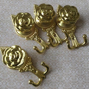 May include: Four decorative gold-coloured metal hooks. Each hook has a rose-shaped design at the top, with detailed petals and leaves. These hooks are suitable for hanging items and add a touch of elegance to any room.