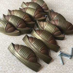 Pode incluir: Um conjunto de puxadores de armário de estilo antigo. Cada puxador apresenta um corpo curvo castanho com riscas verticais douradas e uma moldura dourada. Vários puxadores estão dispostos sobre uma superfície estampada, com parafusos visíveis.