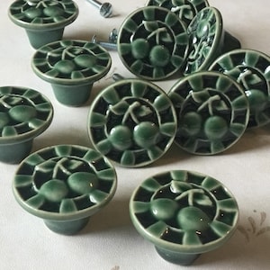 Op de afbeelding: Een verzameling groene keramische kastknoppen met een kersendesign. De knoppen zijn rond met een verhoogd kersmotief en een donkergroene rand. Montageschroeven zijn ook zichtbaar.
