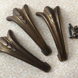 May include: Three antique bronze-coloured furniture legs with a decorative floral design. The legs are curved and have a distressed finish. A small pile of screws is next to the legs, suggesting they are ready for installation.