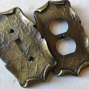May include: Two decorative light switch covers. One has a single switch opening, the other has two outlet openings. Both are textured with a gold and bronze finish, and black screws.