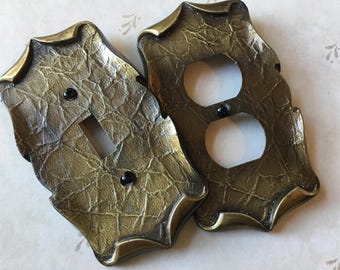 Könnte beinhalten: Zwei dekorative Lichtschalterabdeckungen. Eine hat eine einzelne Schalteröffnung, die andere zwei Steckdosenöffnungen. Beide sind strukturiert mit einer Gold- und Bronzefinish und schwarzen Schrauben.