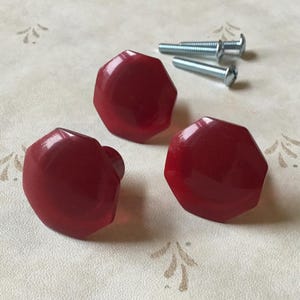 May include: Three faceted, ruby-red cabinet knobs with matching screws. The knobs have a glossy finish and a geometric design. The hardware is set against a cream-coloured background with a subtle floral pattern, suggesting a vintage or antique aesthetic.