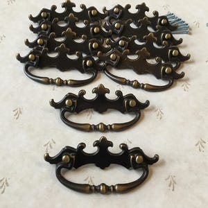 Puede incluir: Un juego de 12 tiradores de cajón de armario de latón antiguo con diseños ornamentados. Los tiradores tienen un diseño decorativo y ornamentado con una manija de bucle.