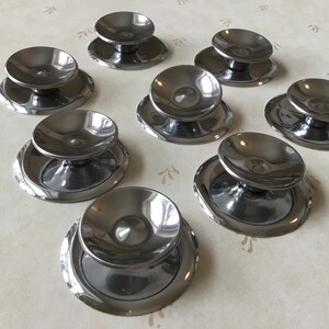 Könnte beinhalten: Acht silberfarbene Metallknöpfe mit einer runden, leicht konkaven Oberseite und einer erhöhten, runden Basis. Die Knöpfe sind in einem Kreis auf einer weißen Oberfläche angeordnet.