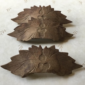 May include: Four intricately carved wooden leaf-shaped decorative pieces. The leaves are a warm brown colour with detailed veining and a central floral design. The pieces are arranged on a light background.