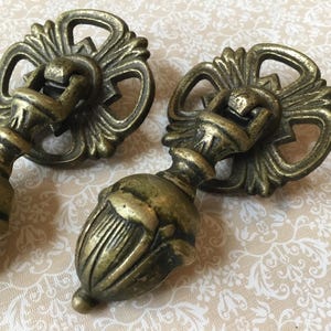 May include: Two antique brass drawer pulls with ornate floral and scrollwork designs. The pulls have a decorative backplate and a teardrop-shaped handle. The brass has a weathered, aged appearance.