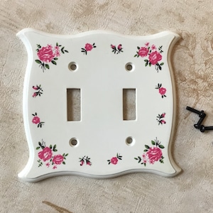 Puede incluir: Cubierta de interruptor de luz doble blanca con un diseño floral de rosas rosas y hojas verdes. La cubierta tiene un borde festoneado e incluye tornillos de montaje. Una adición encantadora a cualquier habitación.