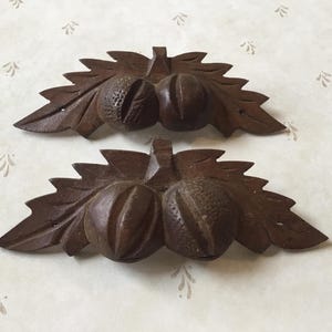 Może przedstawiać: Dwie rzeźbione drewniane dekoracje z żołędziami i liśćmi. Każdy element przedstawia dwa żołędzie i kilka liści, wszystko w ciemnobrązowym kolorze. Rzeźby są szczegółowe i wydają się być w stylu vintage lub antycznym.