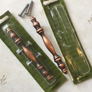 May include: Two antique copper cabinet door pulls with ornate designs. The pulls are packaged on green cards with the brand name "Amerock" printed on them.