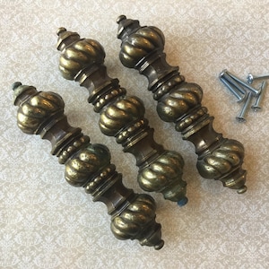 May include: Three antique brass cabinet pulls with an ornate design. Each pull features a twisted, bulbous centre and detailed top and bottom. Several silver screws are also included.