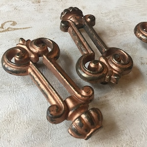 May include: Three antique-style, ornate, copper-toned metal drawer pulls with a decorative scroll design.