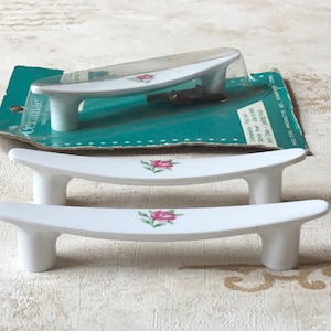 May include: White ceramic cabinet pulls with a curved design and a pink rose decal. One set is still in its original packaging. The pulls have a smooth, glossy finish. The pulls are approximately 13 cm long.