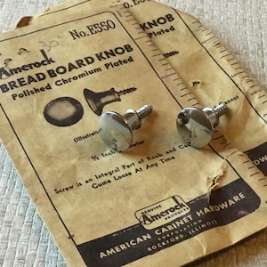 以下が含まれることがあります： 光沢のあるクロム製のパンボードノブ2個とネジのクローズアップ。パッケージには「Amerock Bread Board Knob Polished Chromium Plated No. E550」と「American Cabinet Hardware Corporation Rockford, Illinois」と記載されています。