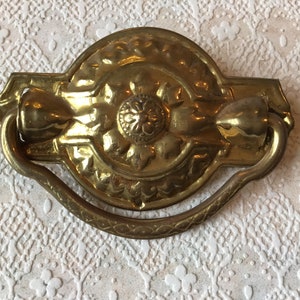 May include: A vintage brass door pull with a decorative floral design. The pull has a loop handle and is attached to a rectangular plate.