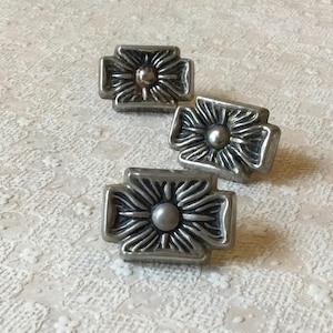 Op de afbeelding: Drie zilveren metalen ladegrepen met een bloemmotief. Elke greep heeft een verhoogde middelste cirkel en een kruisvormige basis.