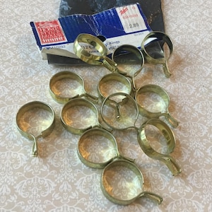 Könnte beinhalten: Eine Sammlung messingfarbener Gardinenringe mit kreisförmigem Design und kleinem Haken. Die Ringe sind auf einer gemusterten Oberfläche angeordnet, mit einem teilweise sichtbaren Pappkarton im Hintergrund.