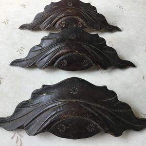 May include: Three dark brown, leaf-shaped decorative wooden appliques. Each applique has a carved floral design in the centre and is set against a patterned background. These appliques are likely for furniture or craft projects.