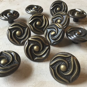 Może przedstawiać: Zestaw 12 antycznych mosiężnych gałek do szafek z ozdobnym wzorem w kształcie spirali. Każda gałka ma wypukły, okrągły środek i fakturowany, spiralny wzór.