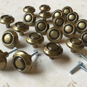 May include: A selection of antique brass cabinet knobs, mushroom-shaped in design. The knobs feature a circular face with a recessed centre and are supplied with silver screws. Ideal for furniture restoration or adding a vintage touch to cabinets.