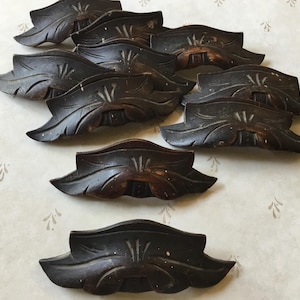 Puede incluir: Un conjunto de elementos decorativos de madera tallada de color marrón oscuro, posiblemente tiradores de cajones o adornos de muebles, dispuestos sobre una superficie estampada. Cada pieza presenta un diseño en forma de hoja con detalles intrincados y un acabado envejecido.