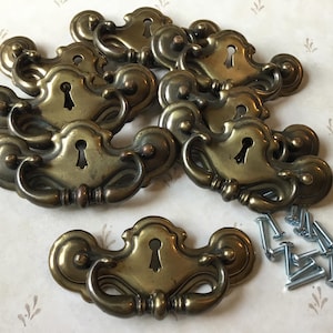 May include: A set of antique brass drawer pulls, featuring keyhole designs and curved handles. The hardware has a warm, aged brass finish, and includes mounting screws. Suitable for vintage furniture restoration.