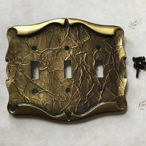 May include: An ornate, antique-style light switch cover with a textured, gold finish. It features three switch openings and decorative edges. Includes black screws for installation.
