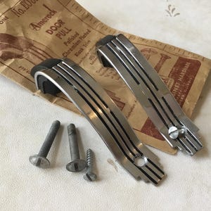 May include: Two polished chrome door pulls with black trim and a decorative design. The pulls are packaged with screws. The packaging includes the text "Amerock Door Pull, Polished Chromium Plated, Black Trim".