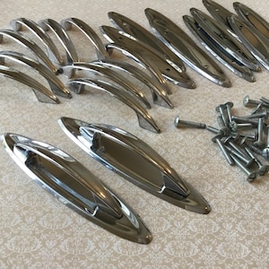 Pode incluir: Uma coleção de ferragens de armário prateadas e brilhantes, incluindo várias alças curvas e puxadores alongados em forma oval. Há também uma pilha de parafusos prateados, sugerindo um conjunto para instalação. As ferragens são provavelmente para móveis ou armários.