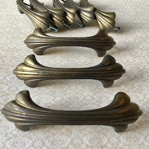 May include: Antique brass-toned cabinet pulls with a decorative, fluted design. The pulls are arranged in a group, showcasing their ornate shape and mounting hardware. The hardware is designed for easy installation on furniture.