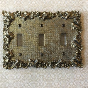 May include: An antique-style light switch plate with three switches. The plate has a textured, metallic finish and is framed with an ornate floral and leaf design. The plate is secured with four screws.