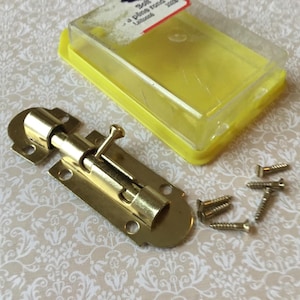 May include: A brass-colored barrel bolt with a sliding mechanism, alongside several small screws and a yellow plastic container. The bolt is designed for securing doors or other closures. The hardware is displayed on a patterned surface.