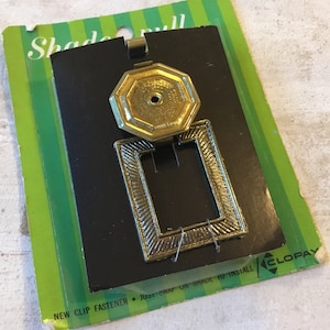 Puede incluir: Tirador de cortina dorado con un marco cuadrado y una parte superior redonda octogonal. El tirador está empaquetado en una tarjeta verde y blanca con el texto "Shade Pull" y "New Clip Fastener - Just Snap On Shade To Install / Clopay".