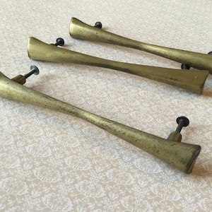 Puede incluir: Cuatro tiradores de armario de latón vintage con un diseño único y alargado. Cada tirador presenta un pomo redondo oscuro y un extremo ligeramente curvado y rectangular. El latón tiene una pátina cálida y envejecida, lo que sugiere un estilo clásico o antiguo.