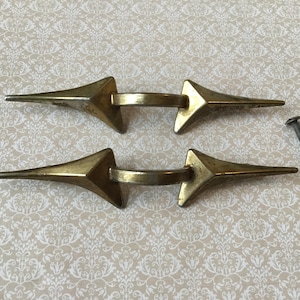 以下が含まれることがあります： 尖った矢印のデザインの、ヴィンテージ真鍮製引き出しプル2つ。各プルには中央のハンドルと2つの尖った端があります。ネジの小さな山が含まれています。プルは模様のある背景にあります。