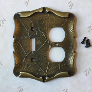 May include: A decorative brass-coloured light switch cover with a textured surface. The cover has a switch opening and two outlet openings. Includes four small black screws for installation. The cover has a decorative, ornate edge.