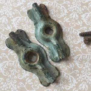 Op de afbeelding: Twee antieke, verweerde, teal-kleurige metalen decoratieve stukken met een centraal gat, vergezeld van twee bruine schroeven. De stukken hebben een unieke, organische vorm en vertonen tekenen van ouderdom en slijtage.