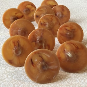 Op de afbeelding: Een verzameling van elf ronde, amberkleurige kastknoppen met een gemarmerd effect. Elke knop heeft een centrale, cirkelvormige inkeping. De knoppen zijn gerangschikt op een patroon, gebroken witte ondergrond.