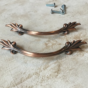 May include: Two antique copper drawer pulls with ornate floral designs. The pulls are curved and have a distressed finish.
