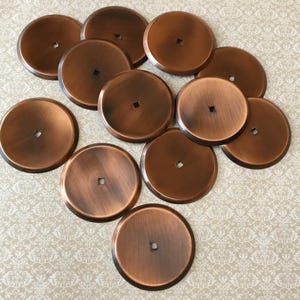 Può includere: Una collezione di undici pomelli per mobili rotondi color rame. Ogni pomello ha un foro centrale quadrato o circolare. I pomelli sono disposti su una superficie chiara a motivi. I pomelli hanno una finitura metallica.