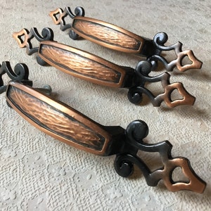 Peut inclure: Trois poignées de tiroir anciennes avec une finition en cuivre martelé et des accents en métal noir. Les poignées ont un design décoratif et orné.