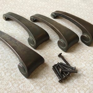May include: Set of five antique bronze-toned cabinet pulls with a curved handle design. Each pull features a decorative end with a circular detail. Includes a pile of matching screws for installation.