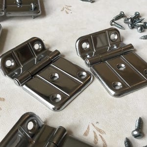 Puede incluir: Dos bisagras de metal plateadas con acabado pulido. Las bisagras están cerradas y tienen forma rectangular. Las bisagras están sobre una superficie blanca con pequeños tornillos plateados esparcidos a su alrededor.