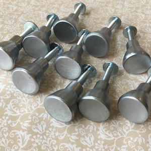 May include: Ten silver-coloured metal cabinet knobs with a threaded bolt and a rounded, flat face. The knobs are arranged on a patterned beige surface, highlighting their simple, industrial design.