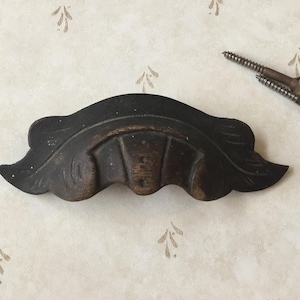 May include: A dark brown, decorative wooden drawer pull with a curved shape and carved patterns. Two screws are placed nearby. The drawer pull is likely vintage or antique.