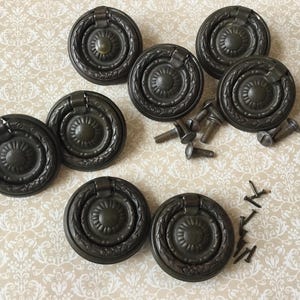 Op de afbeelding: Een verzameling van acht antieke bronzen ringtrekkers met decoratieve bloemendetails. Inclusief verschillende schroeven voor installatie. Perfect voor het restaureren van meubels of het toevoegen van een vintage touch aan kasten en lades.