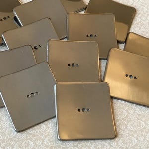 May include: A collection of square, metallic coasters with a brushed finish. Each coaster has three small, evenly spaced holes. The coasters are in varying shades of silver and gold, reflecting light.